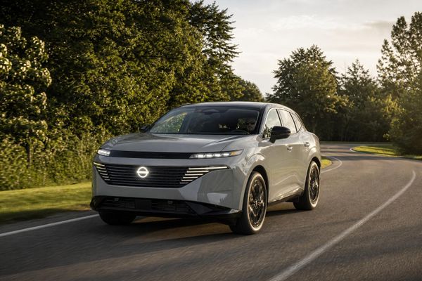 Frontal trekvarts-vy av en ljusgrå 2027 Nissan Murano som kör på en slingrande asfaltväg omgiven av gröna träd i mjukt dagsljus.