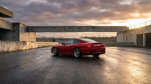 En röd Ferrari 550 Maranello parkerad på en våt asfaltbana framför en brutalistisk betongbyggnad i solnedgången.