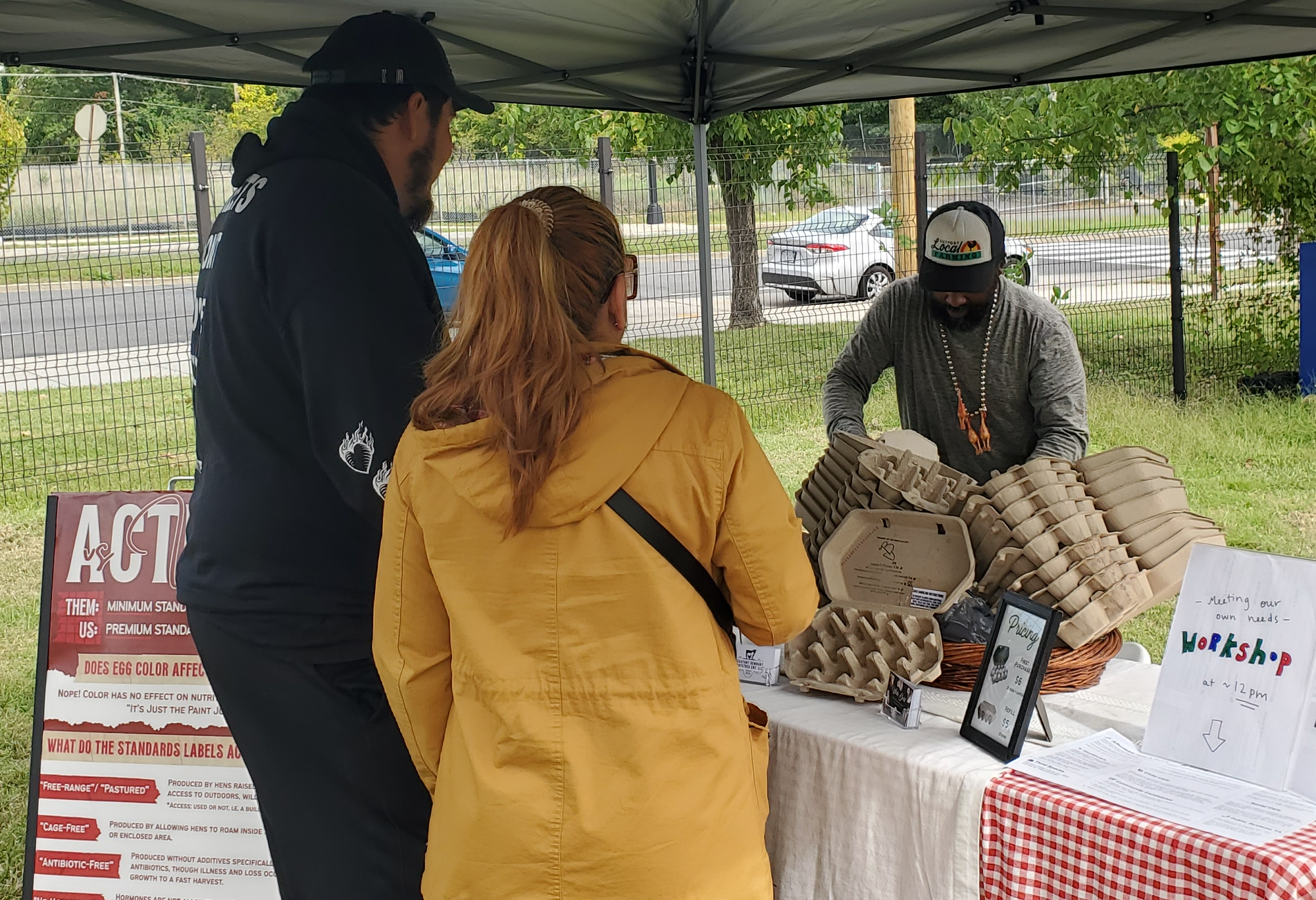 A couple in a black sweatshirt and a yellow jacket get eggs from a farmer table under a tent, surrounded by grass