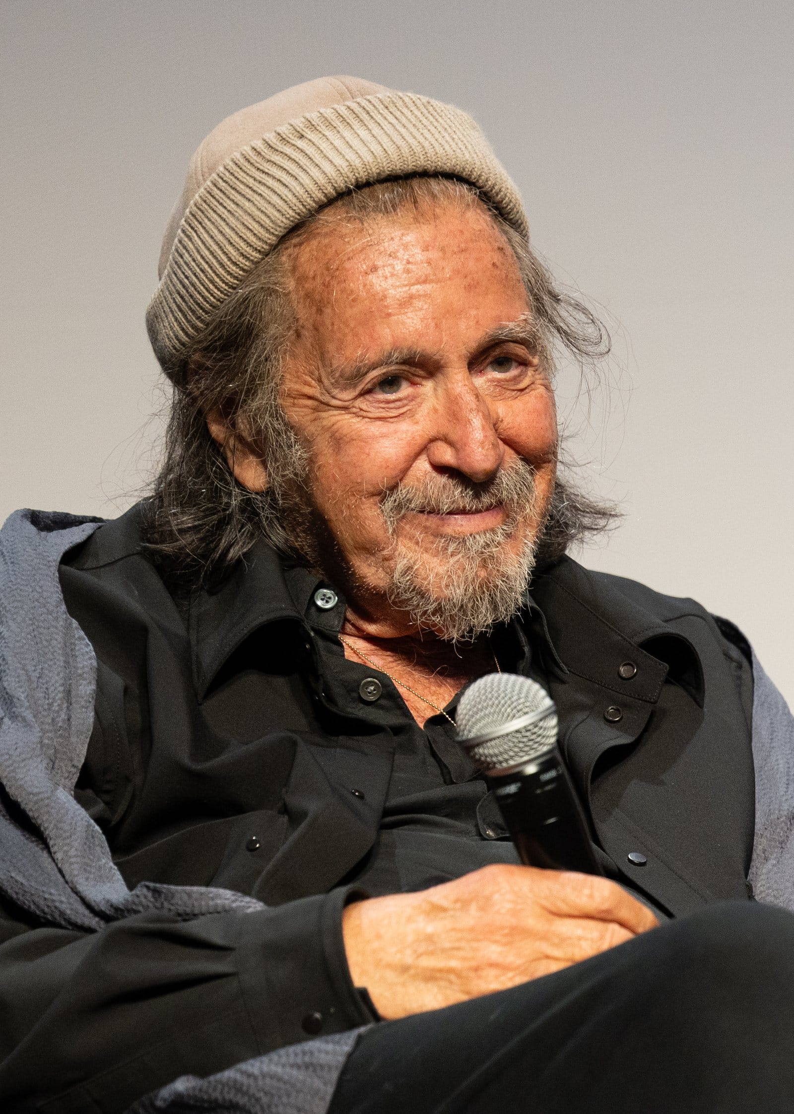 Al Pacino in a knit cap smiles while holding a microphone at a Tribeca Q&A