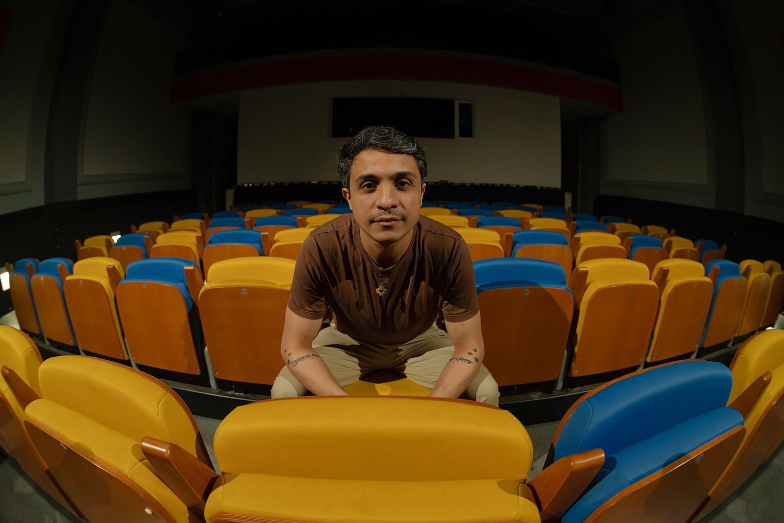 Lorent Saleh seated alone among rows of yellow and blue theater seats