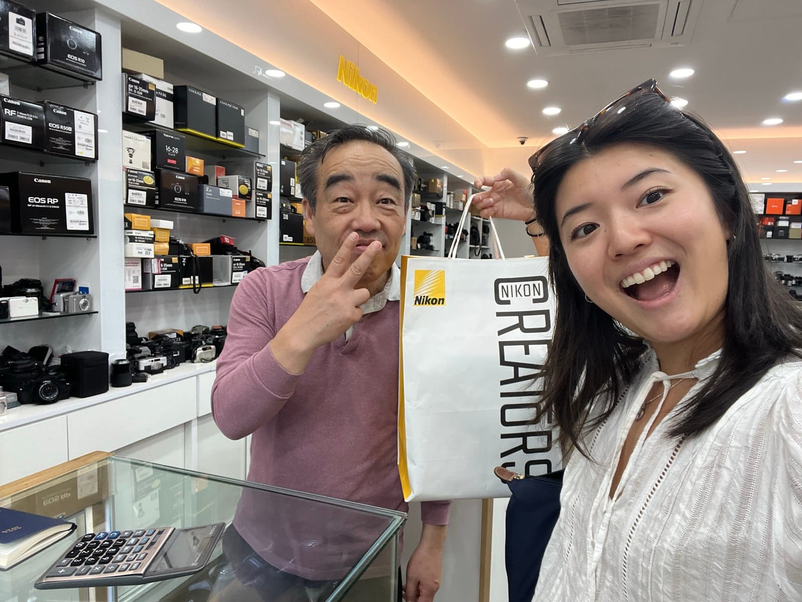 Sara Komatsu takes a selfie with a camera store owner, holding up a shopping bag