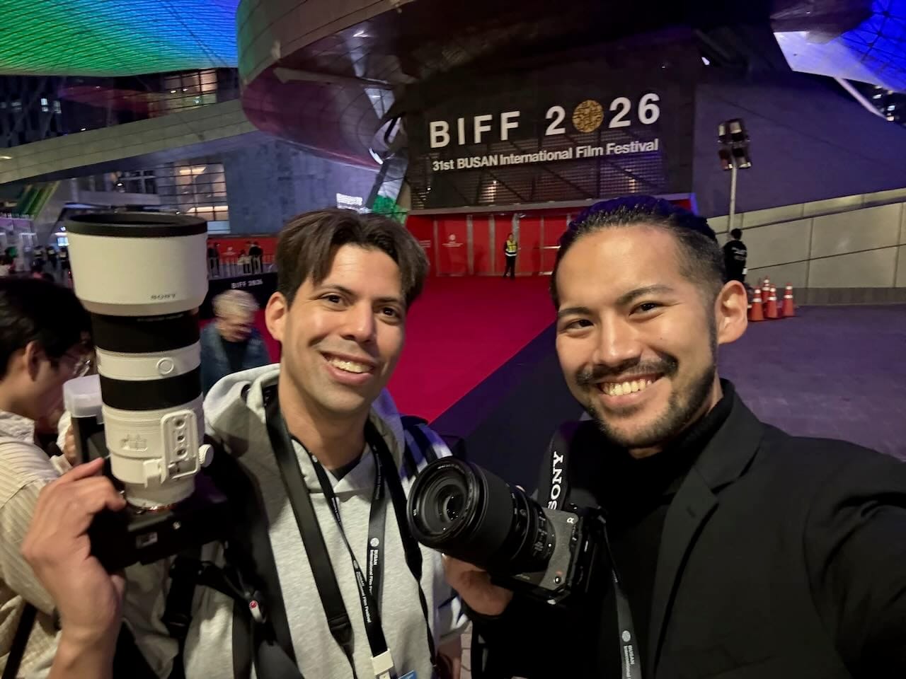 Omar Breeveld and Daiei Onoguchi smile with their cameras in front of the BIFF 2026 red carpet