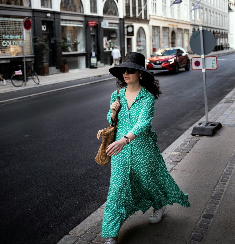 A woman walking along a quiet city street wearing a long green patterned dress and white sneakers, plus a wicker sun hat, and carrying a wicker shoulder bag.