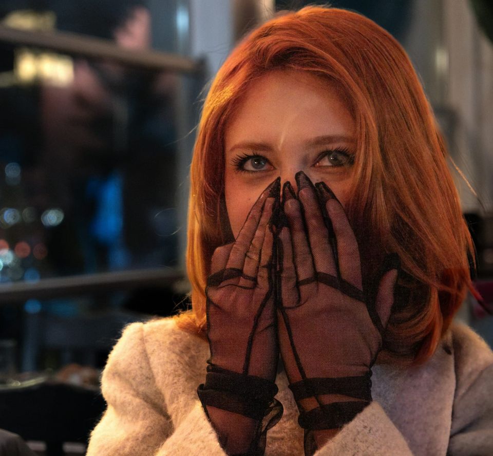 A red-haired young woman wearing a wool coat hides the lower half of her face with sheer-gloved hands
