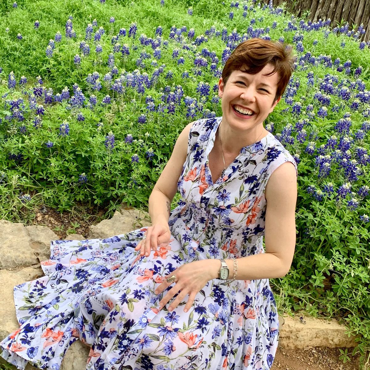 A white woman with short auburn hair, wearing a floral dress and silver jewelry, grins broadly up from her seat on the stone edge of a flowerbed filled with Texas Bluebonnets in peak bloom.