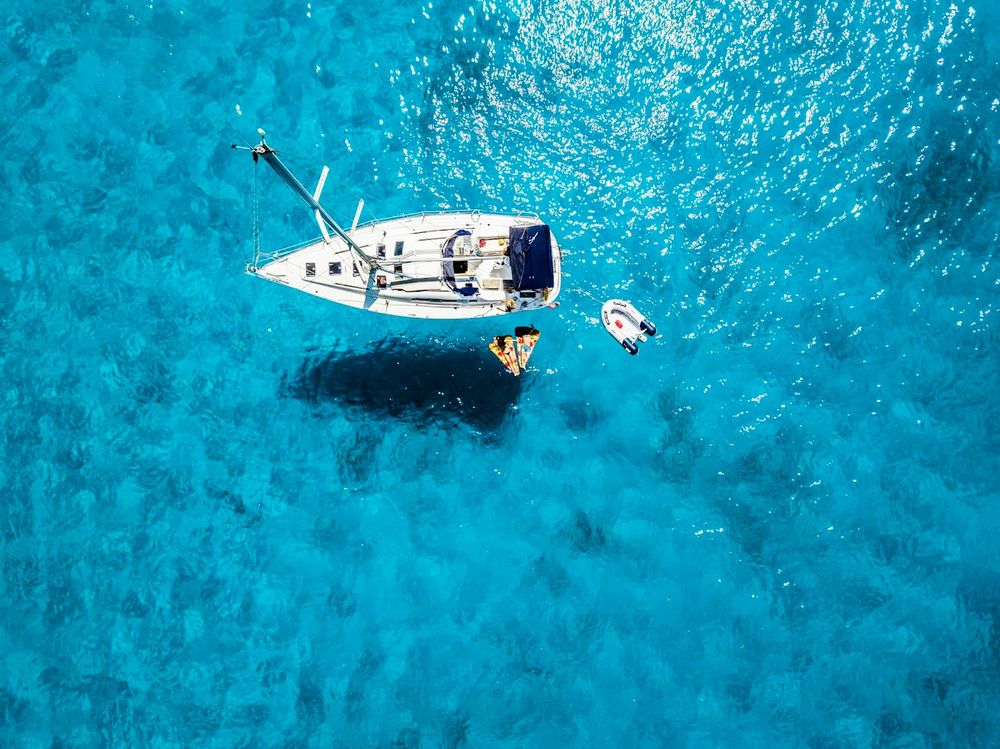 Aerial view of a single white-hulled sailboat at anchor in shallow turquoise water, with a small tender tied alongside