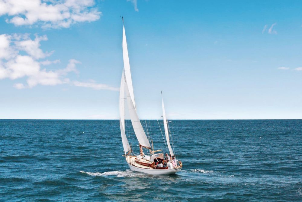 A small white sloop under sail with a crew visible aboard on the open ocean with an empty horizon behind