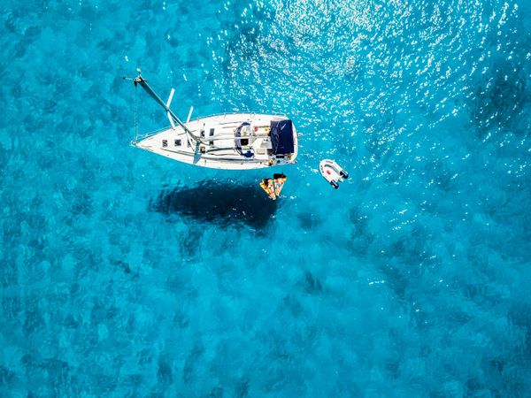 Aerial view of a single white-hulled sailboat at anchor in shallow turquoise water, with a small tender tied alongside