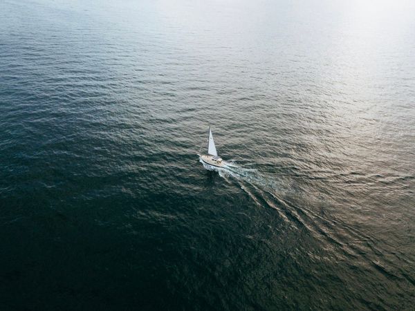 Aerial view of a sailboat underway alone on open ocean water with sun glare on the surface