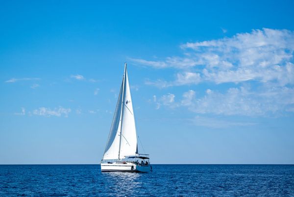 A white cruising catamaran under full main and genoa on deep blue open water beneath a clear sky with scattered clouds