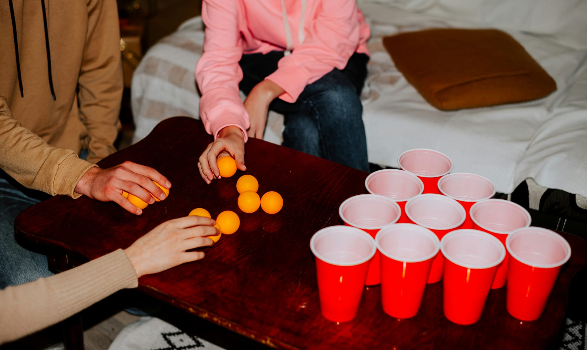 Tre personer spelar beer pong med orange pingisbollar och röda plastmuggar på ett bord i ett vardagsrum.