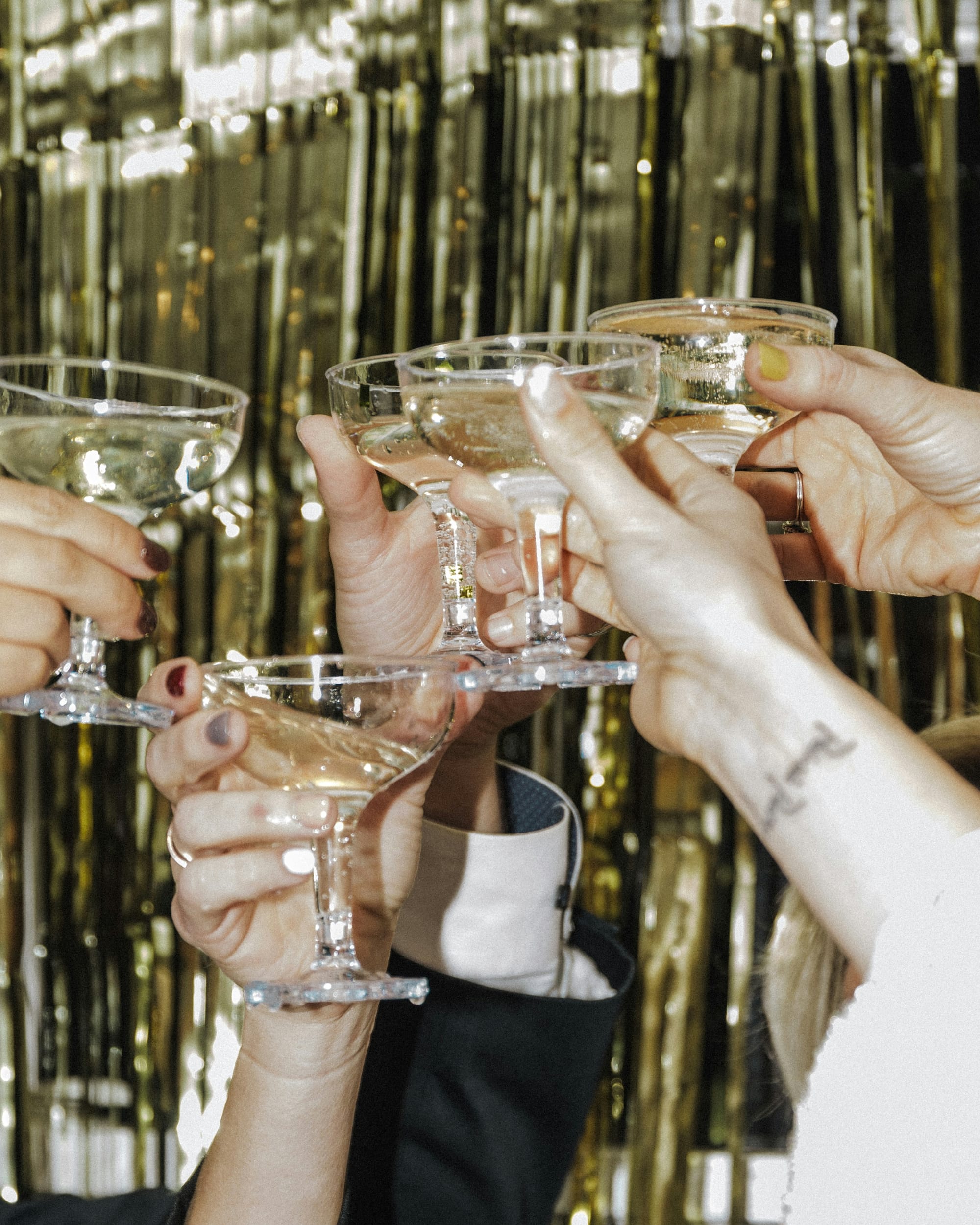 Flera händer som skålar med champagneglas framför ett glittrande guldigt draperi under en nyårsfest.