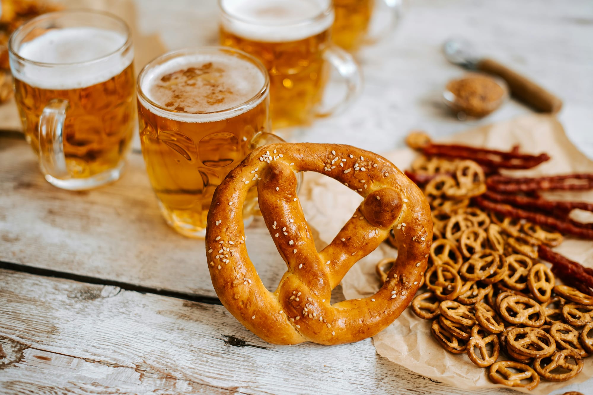 En stor, färsk pretzel med sesamfrön ligger i förgrunden, omgiven av mindre kringlor och ölglas fyllda med oktoberfest öl, placerade på ett rustikt träbord. Bilden visar klassisk oktoberfest mat.