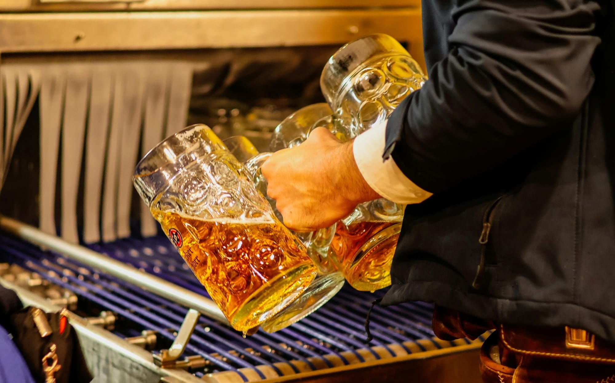 En person i lederhosen håller två stora ölsejdlar fyllda med skummande öl, redo att serveras under en oktoberfest. Sejdlarna är fyllda från en bardisk med kranar i bakgrunden.