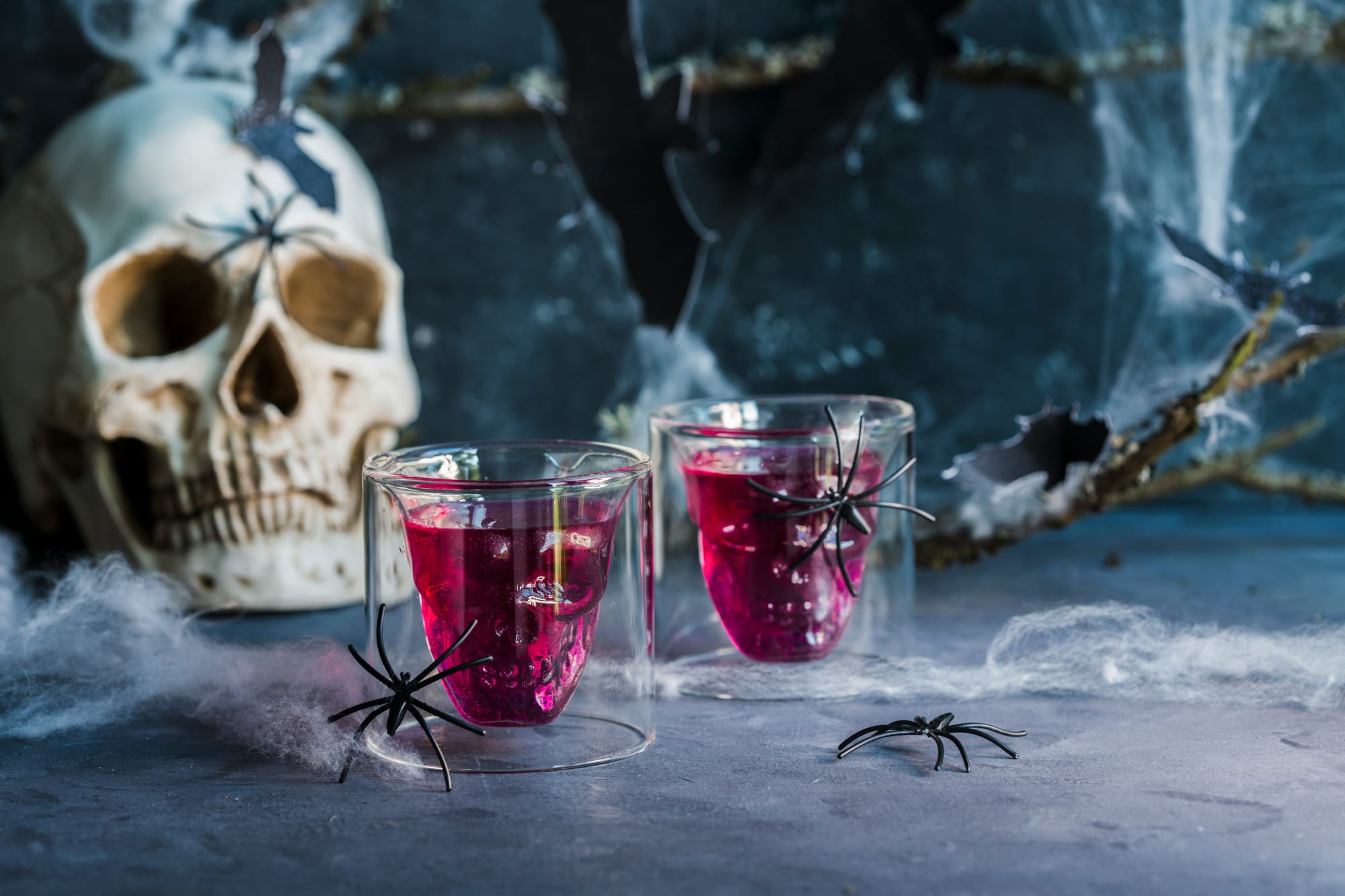 Två glas med röd dryck och plastspindlar framför en dekorerad Halloween-skalle och spindelnät, perfekt för Halloween fest drinkar.