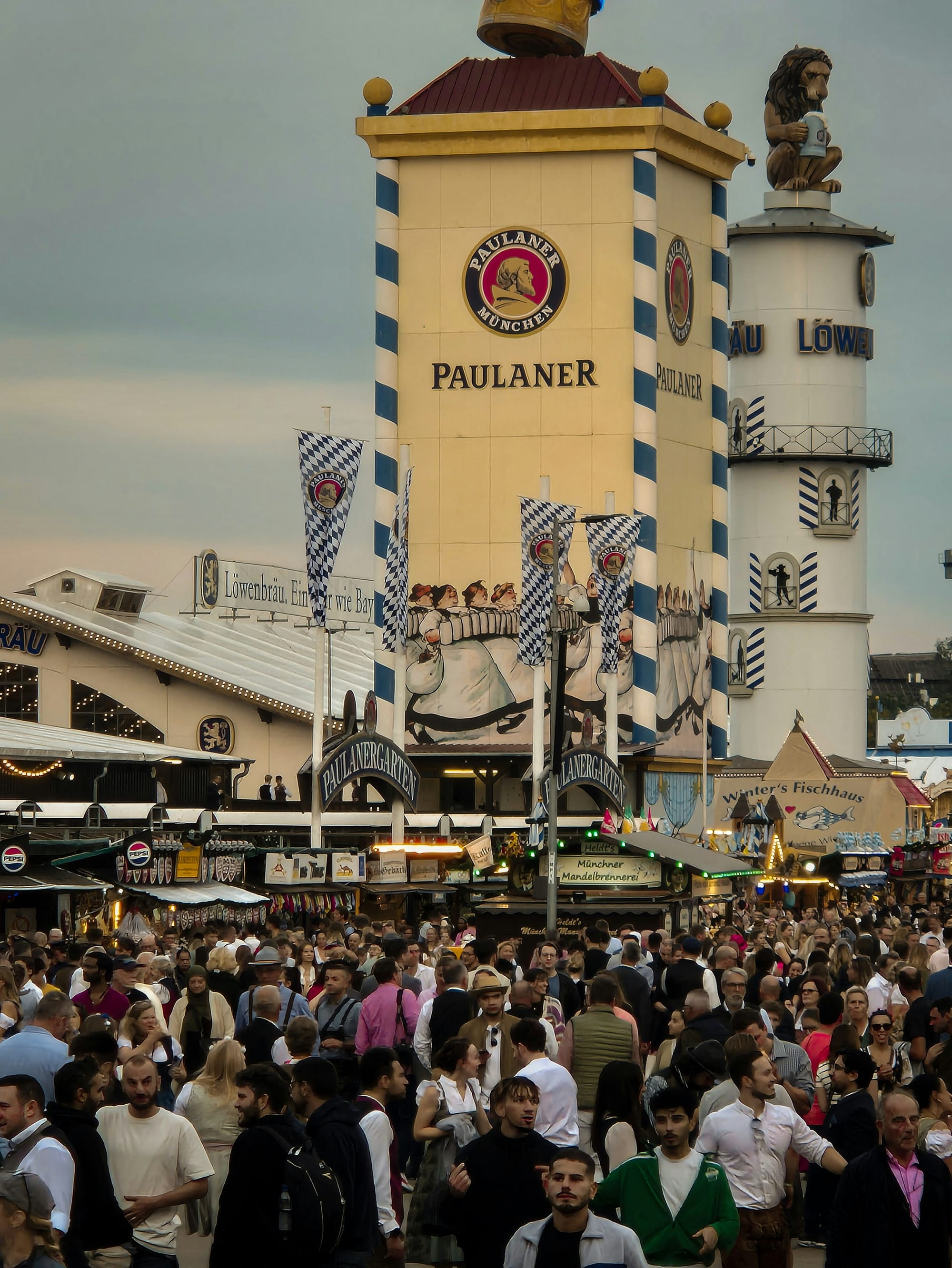 En livlig folkmassa minglar utanför en stor Paulaner tältbyggnad på en oktoberfest. Området är fyllt med människor och byggnader dekorerade i bayerska färger.