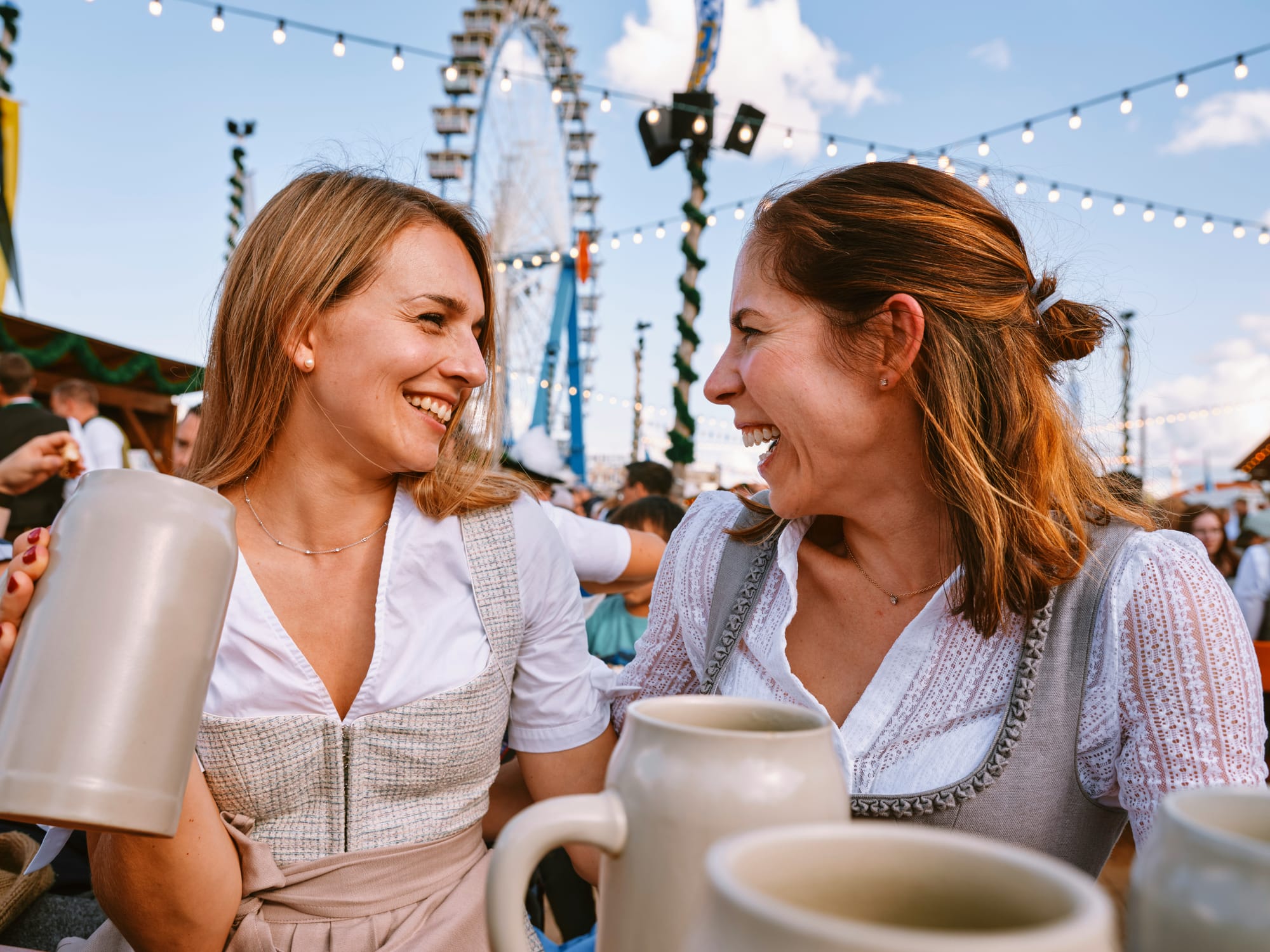 Två glada kvinnor i traditionella dirndl kläder delar ett skratt vid ett oktoberfest bord, med ett par stora ölsejdlar i förgrunden och ett pariserhjul synligt i bakgrunden. Bilden fångar den livliga stämningen under oktoberfest.