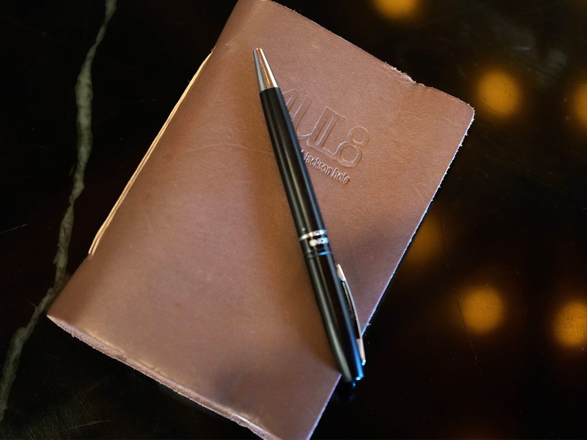 Embossed 4mul8 Jackson Hole leather journal with a pen resting on top, warm lighting blurred behind.