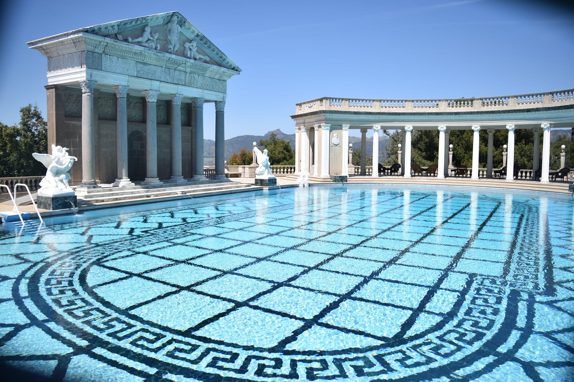 Wide angle shot of William Randolph Hearst's extravagantly ornate pool with mountains and trees on the horizon, very wide and long clear blue water with a black grid along the bottom of the pool bordered by roman style patterns in repeating tile-like shapes. A semicircle columned roof is across at the far end of the long pool, and the narrower side has its own columned roofed structure with carved frescoes on the front and statues of griffins in front at the edge of the pool.