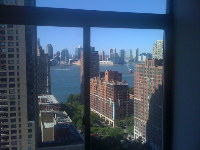 View through apartment windows overlooking Lower Manhattan buildings, green trees, the Hudson River, and the Jersey City skyline under a clear blue sky.