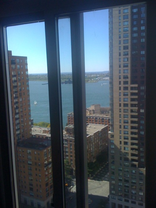 High-angle window view framed between high-rise residential towers looking out over the Hudson River and harbor with sailboats.
