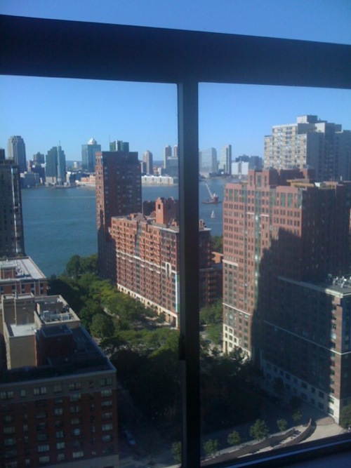 Apartment window perspective showing brick residential buildings, street trees, and the waterfront with the New Jersey skyline across the river.