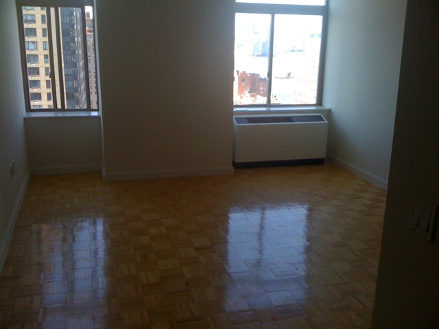 Empty apartment living room with parquet wood floors, white walls, two large windows, and a white climate control radiator unit.