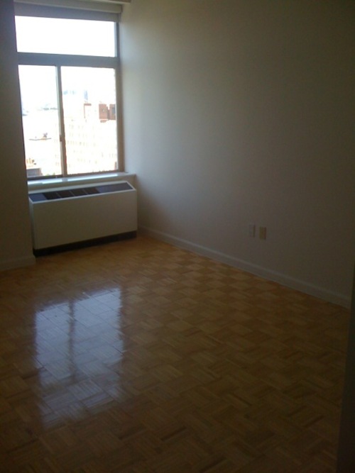 Empty bedroom or living space with shiny parquet wood flooring, white walls, and a large sunlit window above a radiator unit.