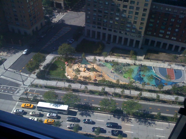 High-angle aerial view looking straight down from the apartment onto West Street traffic, crosswalks, bike paths, and a colorful neighborhood playground with basketball courts.