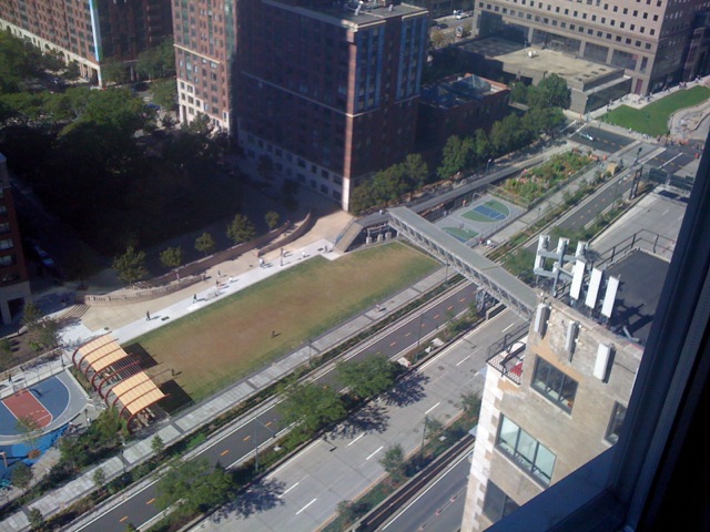 High-angle view from the apartment looking down over a pedestrian overpass bridge, multi-lane roadway, green sports field, and adjacent Battery Park City buildings.