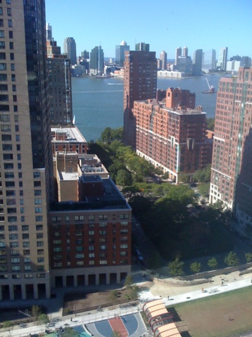 High-angle perspective looking down past apartment towers toward green park trees, athletic courts, the river, and distant cityscape.