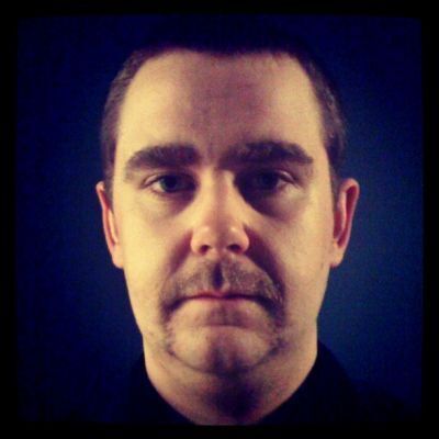 Portrait of a man with a dark brown horseshoe mustache against a deep blue background.