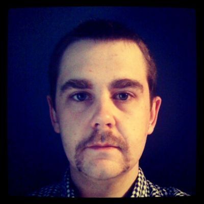 Man with a thick horseshoe mustache wearing a navy gingham shirt against a dark background.
