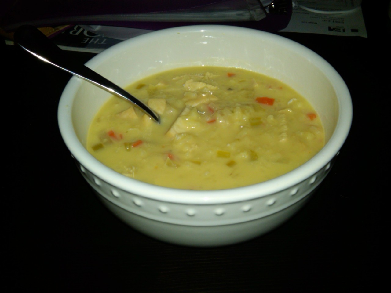 A warm white ceramic bowl filled with homemade chicken and rice soup with carrots and celery, with a metal spoon resting in the broth.