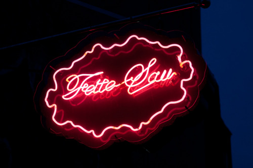 Glowing red cursive neon sign in a scalloped frame reading 'Fette Sau' against the dark evening sky.