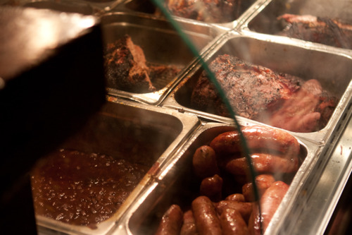Steam rising from steel food trays filled with barbecue meats, sausages, and beans at Fette Sau.