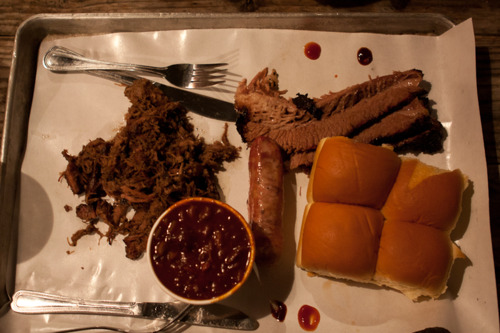 Metal tray with pulled pork, sliced brisket, smoked sausage link, baked beans, and potato rolls.