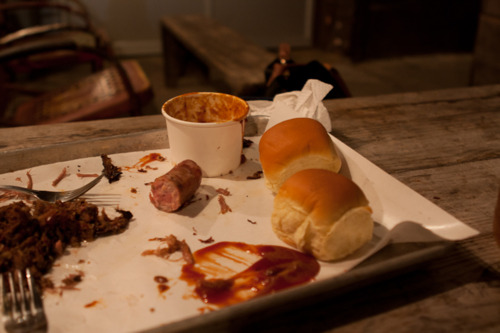 Barbecue platter with leftover pulled pork, sausage, sauce smears, and rolls on a picnic table.