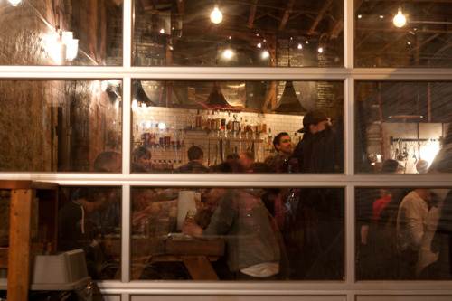 Gridded industrial glass windows looking into the warm, bustling dining room of Fette Sau at night.