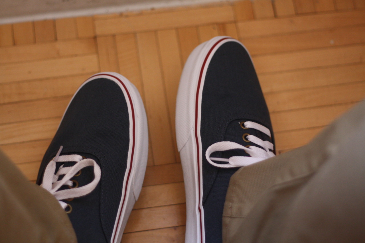 Feet wearing navy blue Vans canvas sneakers with white laces on light wood parquet flooring.