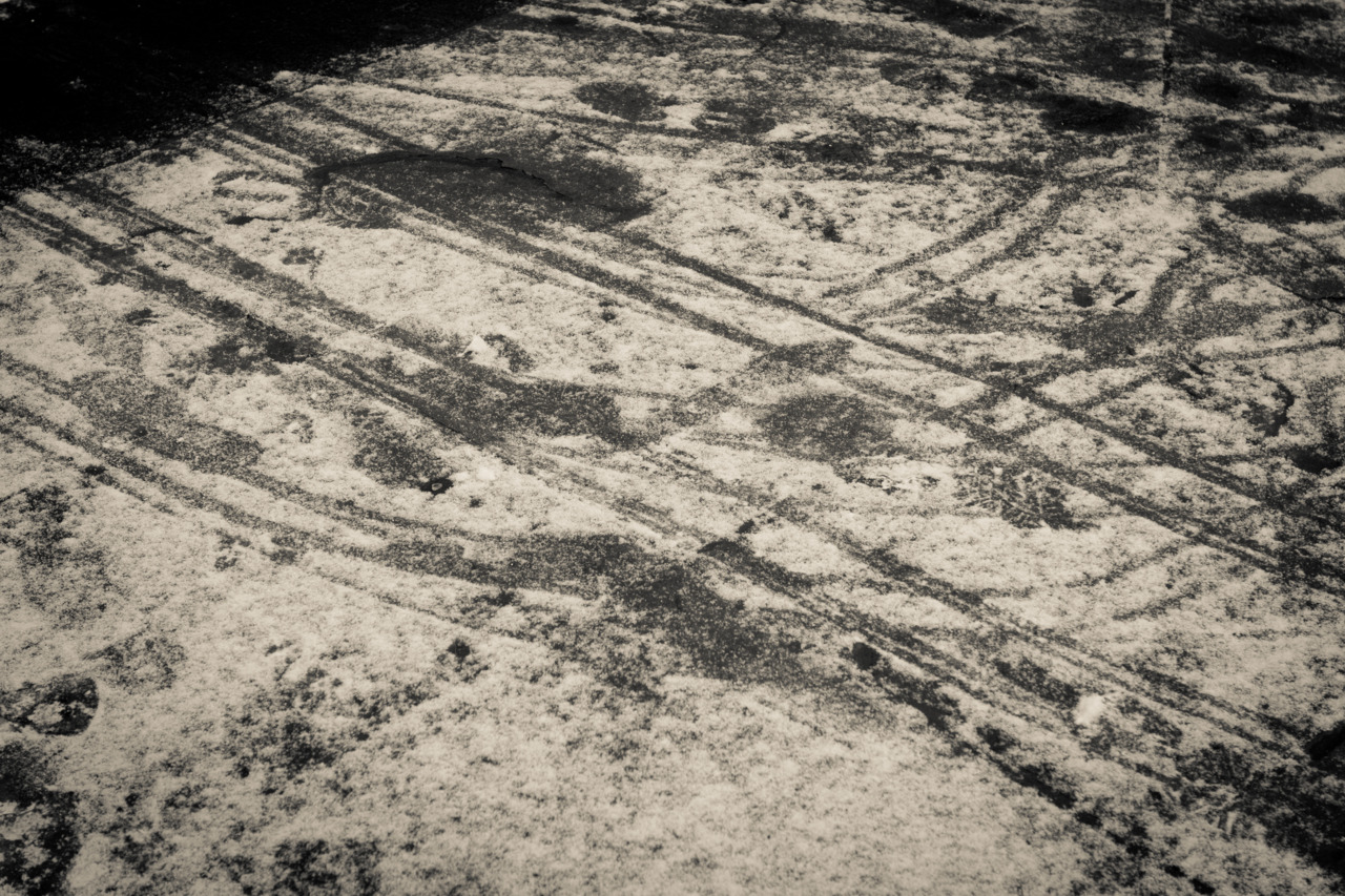 Fresh light snow on asphalt pavement marked with tire tracks, footprints, and dark wet patches.