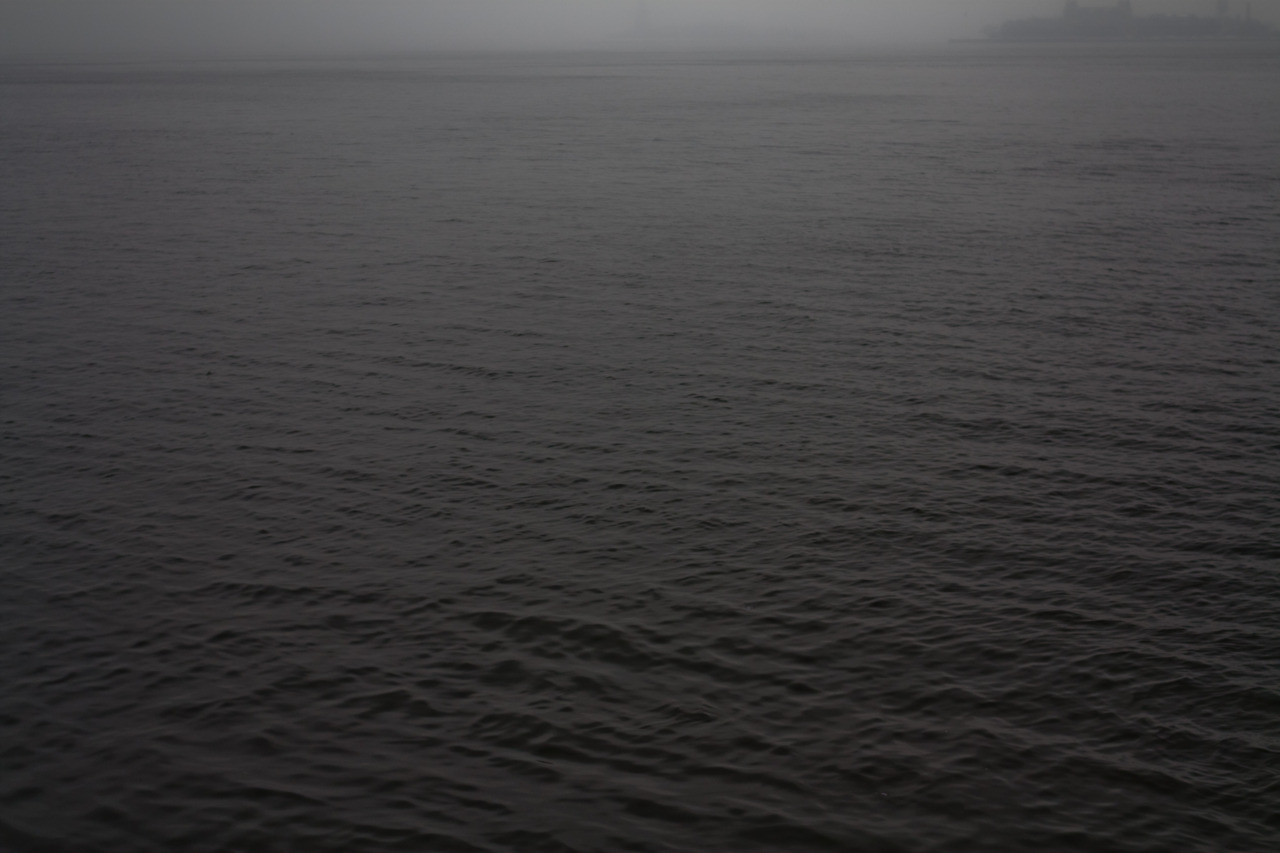 Dark choppy harbor water shrouded in dense morning fog with distant shore silhouettes on the horizon.