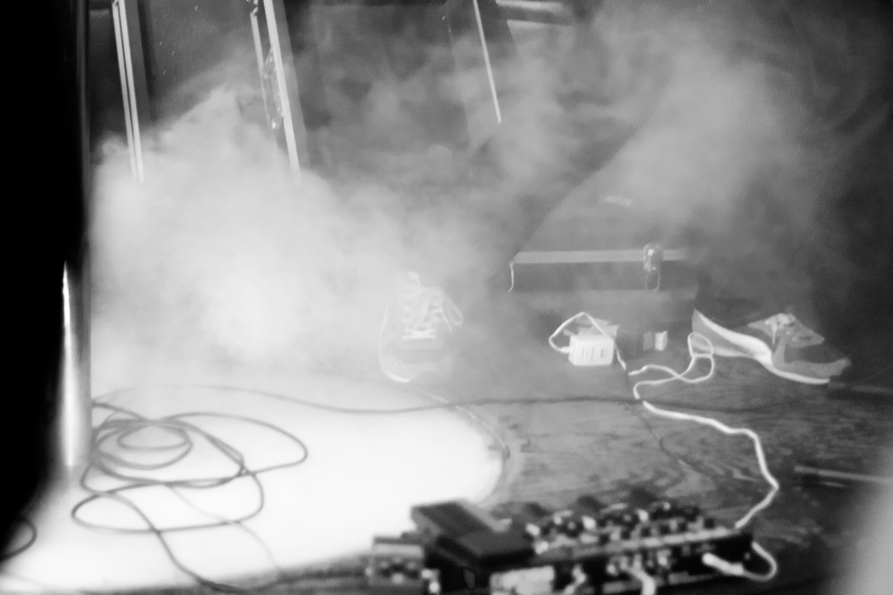 Swirling theatrical haze and spotlight glare over an effects pedal board and audio cables at a concert.