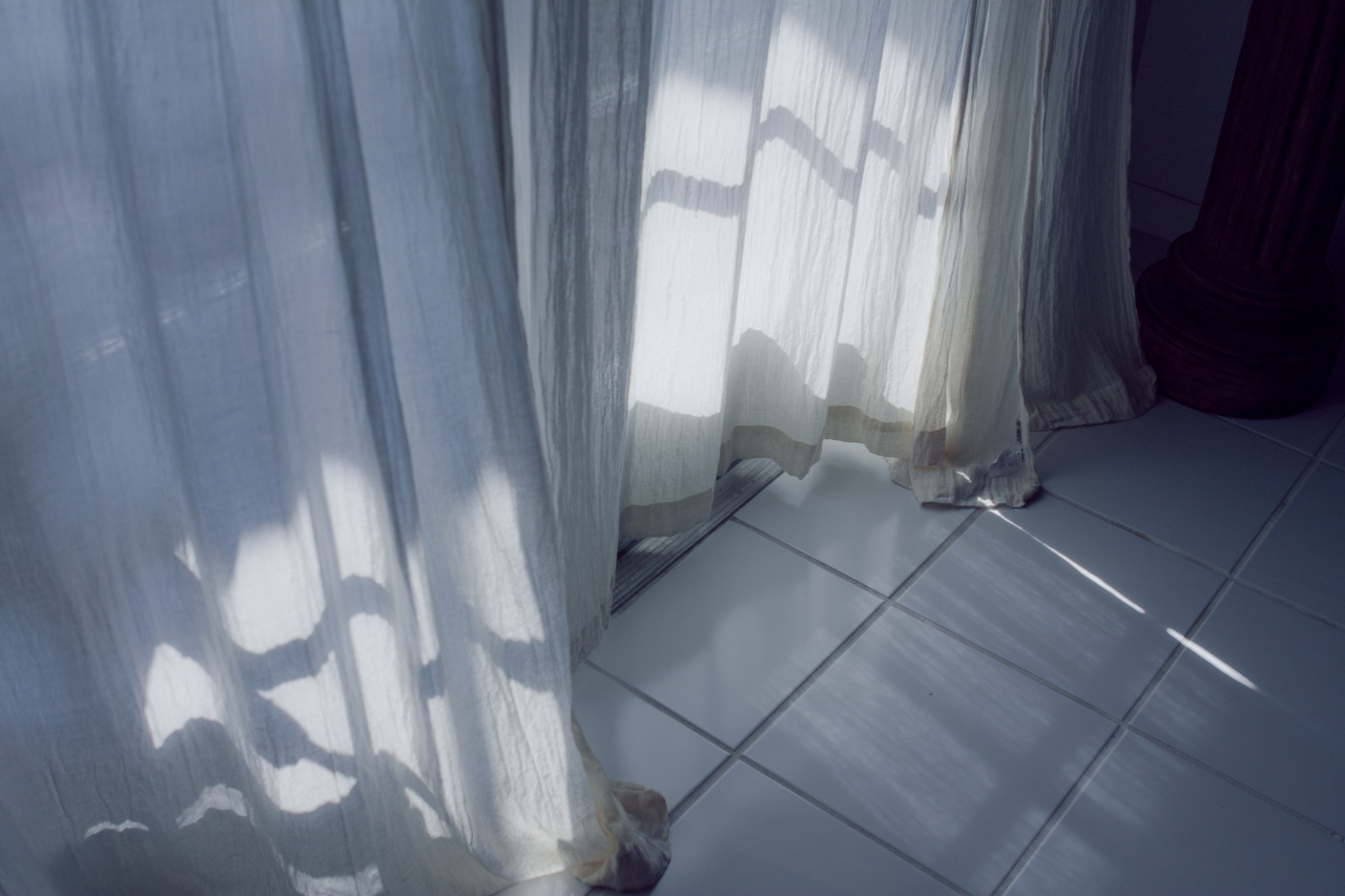 Sunlight through sheer white curtains casting striped shadows across white floor tiles.