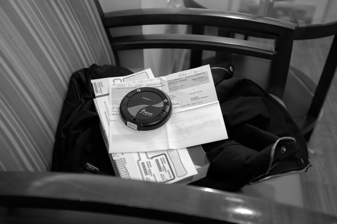 Black and white close-up of a hospital patient pager buzzer resting on medical appointment paperwork atop a chair.