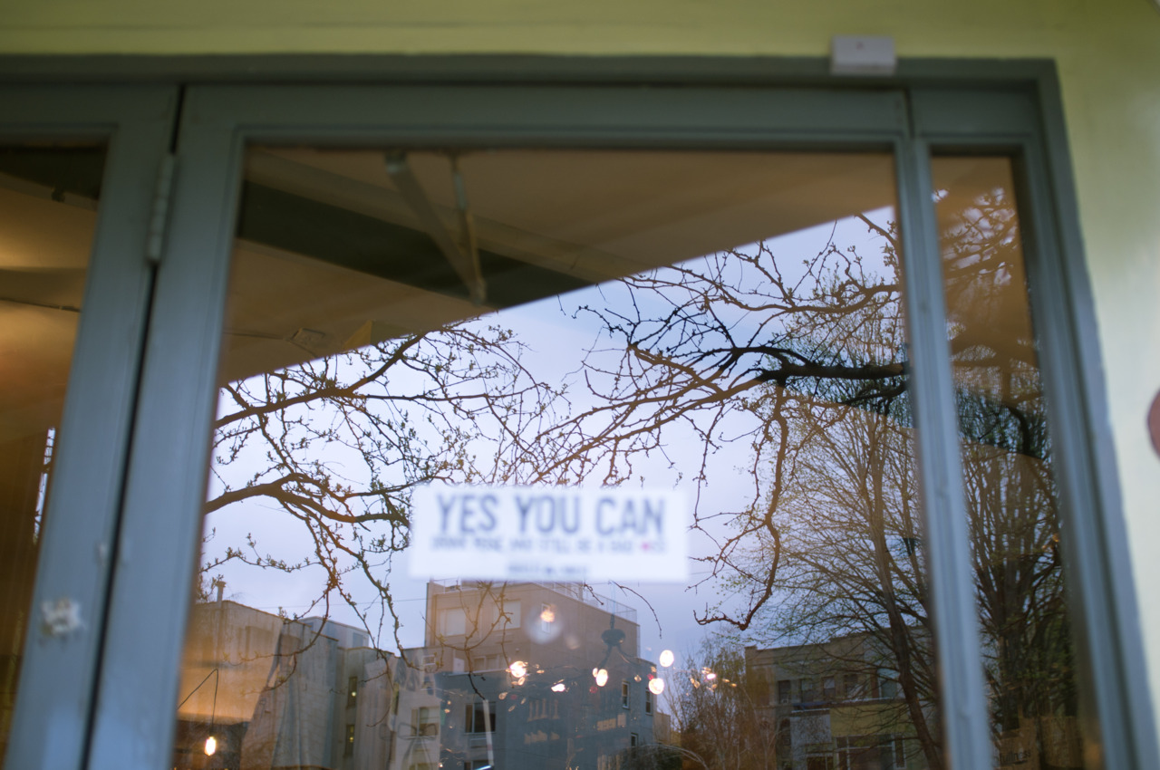 Glass storefront door reflecting bare spring branches and twilight sky with a wine sticker on the pane.