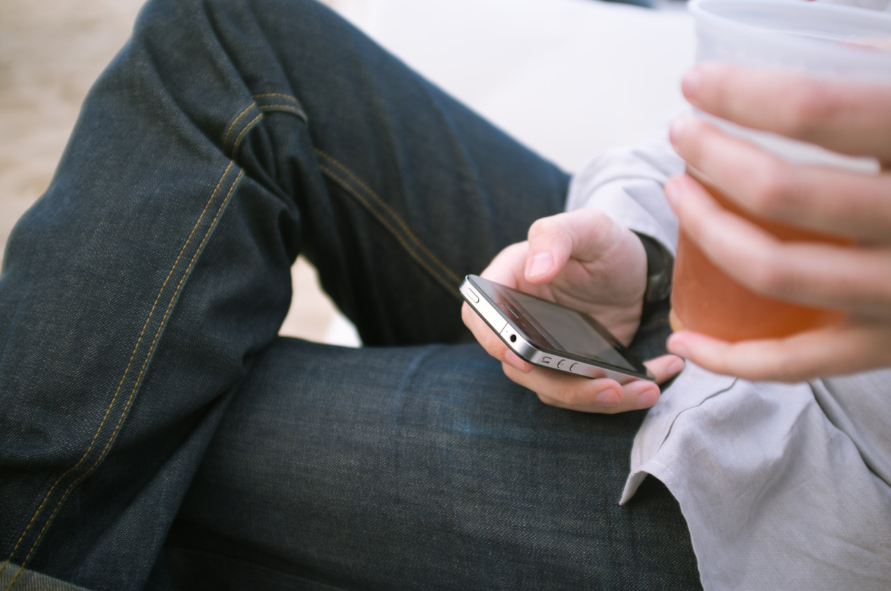 Close-up of a person in dark selvedge denim jeans holding an iPhone 4S in one hand and a cold drink in the other.