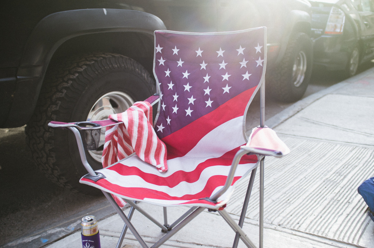 American flag pattern folding chair unfolded on a city sidewalk beside a parked pickup truck in the sun.