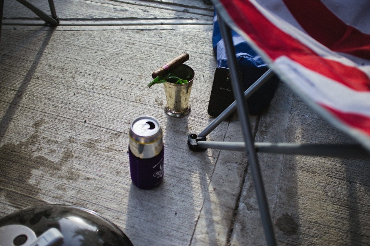 Silver mint julep cup with a lit cigar on its rim beside a beer can and smartphone on an outdoor table.